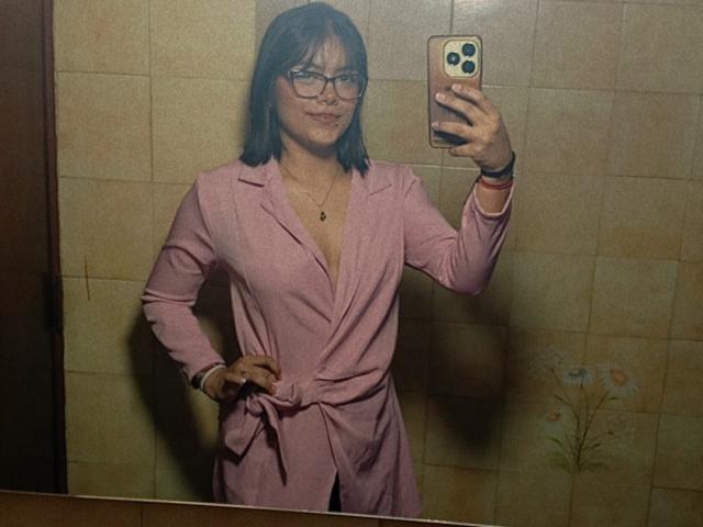 EmilyRoberts with black hair, glasses, and pink robe, standing in bathroom, taking selfie with phone, hand on hip, visible tits, subtle smile, necklace, 1980s style.
