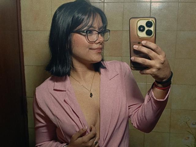 EmilyRoberts with black glasses, pink jacket, and necklace, holding phone, showing her tits, smiling, mirror selfie, beige tiled wall, short black hair, red wristbands.