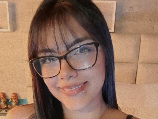 EmilyRobeerts has black glasses, straight black hair with bangs, and a small droplet of saliva on her lower lip. She's smiling slightly. Her green eyes are visible.