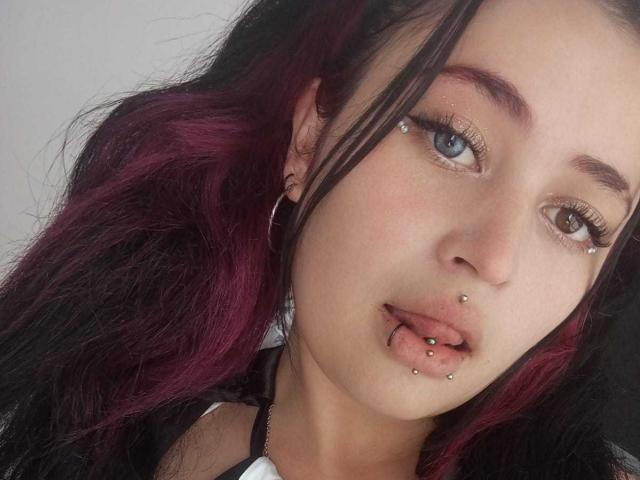 EvaMiller69, con cabello de color púrpura oscuro y negro, ojos azules, múltiples perforaciones faciales, incluyendo en la nariz, el labio inferior y la ceja, y con aros de aro negros..