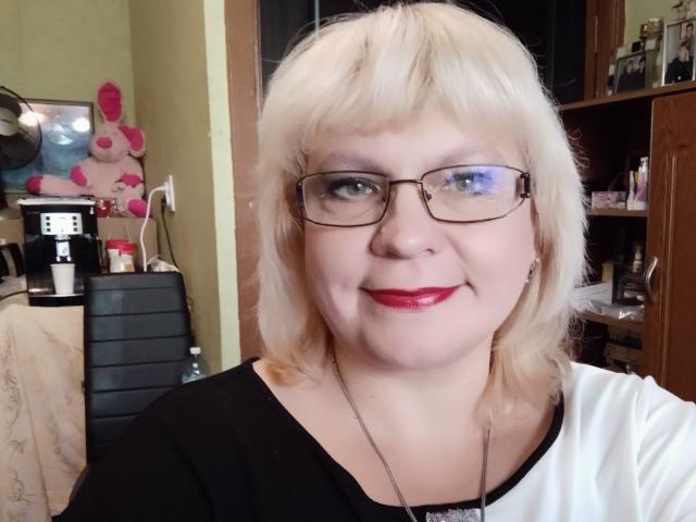 BeverlyCynia has blonde hair, glasses, red lipstick, black and white shirt, and a smiling face. She's a chubby woman with fair skin and visible double chin.