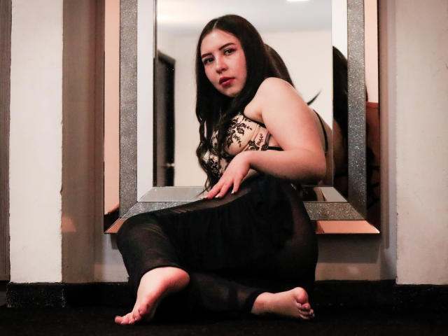MaddieMooree with long black hair, wearing a black lace bra and sheer black dress, sitting barefoot in front of a mirror, looking seductively at the camera.