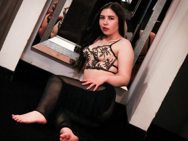 MaddieMooree with long black hair, pale skin, wearing a black lace bra and sheer black skirt, sitting on a black carpet in front of a mirror, looking at camera.