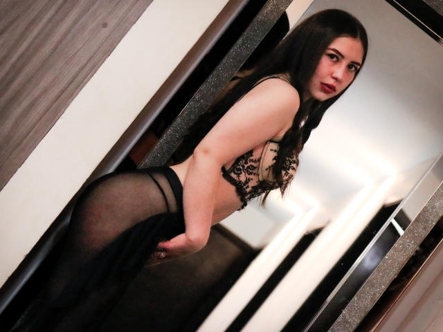 MaddieMooree with long dark hair, wearing a lace bra and sheer black panties, looking over her shoulder, showing her ass and black lace bra. Her hand is on her butt.