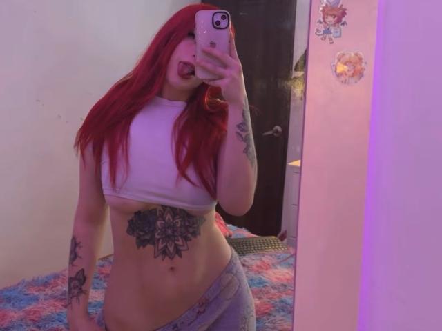 SammyViolet with red hair, tongue out, white crop top, gray pants, tattoos on stomach and arms, holding phone in mirror, showing her tits and ass. Bedroom, purple light.