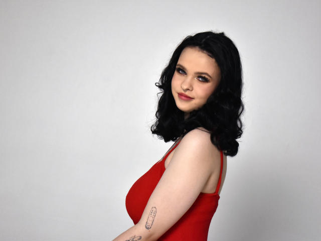 HeliaPolonius with black curly hair, red dress, and tattoo on right arm, smiling at camera, showing her tits and ass, against plain white background.