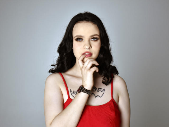 HeliaPolonius, con cabello negro y ondulado, un top rojo y un tatuaje sobre los pezones, mordiéndose el dedo, llevando una pulsera negra, mirando seductoramente a la cámara..