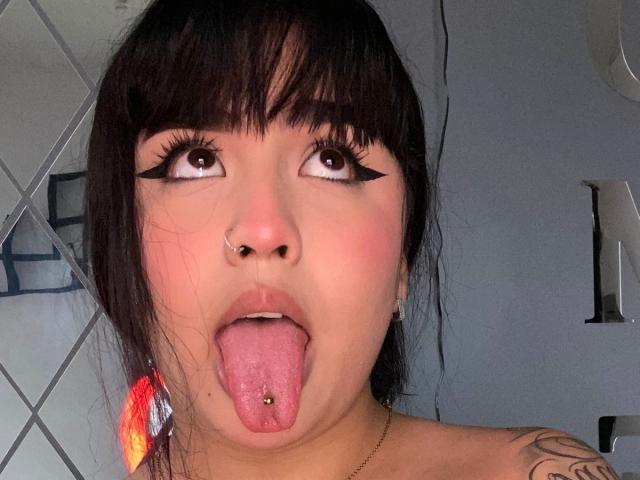 MiaHadid with black eyeliner, nose ring, tongue out, and tongue piercing, looking up with her tongue sticking out, showing her pierced tongue and tattoos.