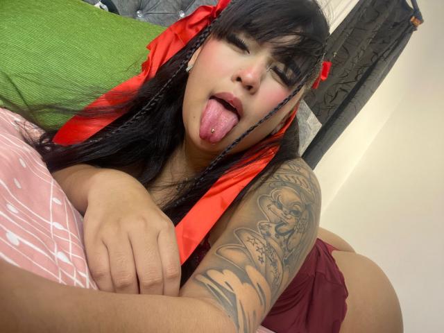 MiaHadid with black hair, red braids, and tongue out, showing tattooed arm, lying on bed, green pillow, pink bedsheet, and red shorts, looking playful and sexy.