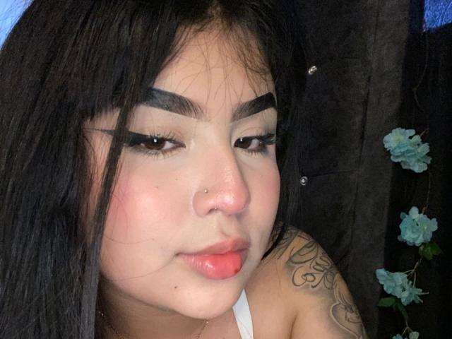 MiaHadid has black hair, thick eyebrows, long eyelashes, a nose piercing, pink lips, and a tattoo on her right shoulder. She's looking slightly down with a subtle smile.