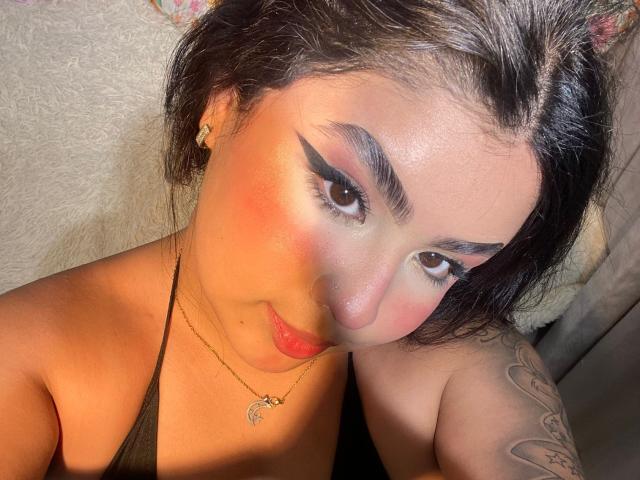 MiaHadid with dark hair, black eyeliner, pink blush, red lipstick, gold necklace, small earrings, tattooed arm, and black halter top, looking at camera with a slight smile.