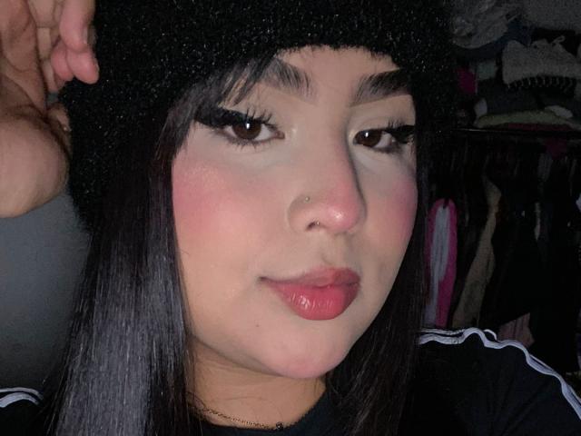 MiaHadid with black wool hat, brown eyes, long black eyelashes, pink blush, nose ring, shiny red lips, and black sports shirt, holding hat with right hand.