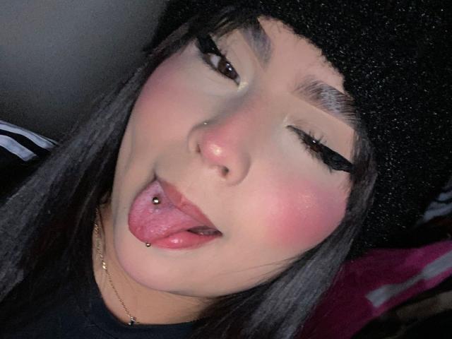 MiaHadid with black beanie, long straight hair, and pink blush, tongue out, showing pierced nose and tongue, heavy black eyeliner, and gold necklace.