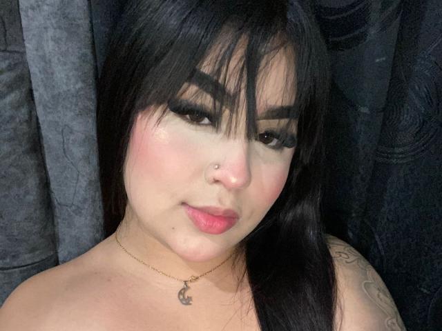 MiaHadid with black hair, thick eyebrows, long eyelashes, pink blush, nose ring, gold necklace, and tattoo on right shoulder, looking at camera with pink lips, topless.
