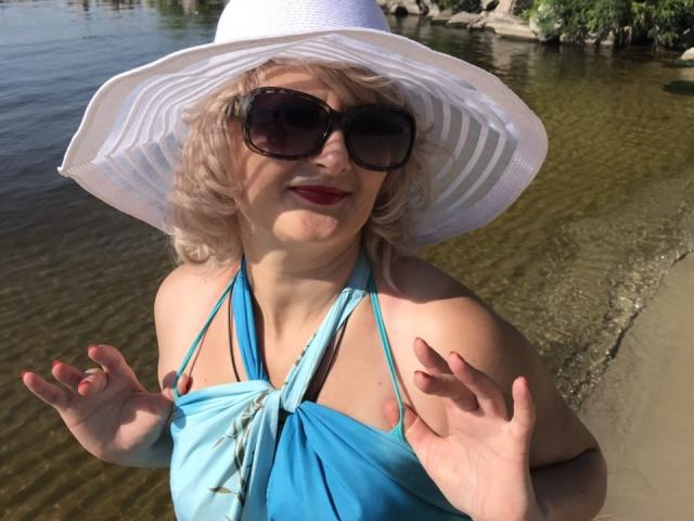NikolinaBounty with curly blonde hair, wearing a white sunhat, black sunglasses, red lipstick, and a blue halter top, standing by a clear water body. Boobs visible under top.