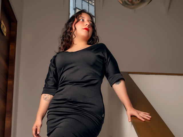 ChloeVelvet with curly black hair, red lipstick, black dress, slightly chubby, tattoo on right arm, leaning on staircase, looking up, sunlight, wooden door, white wall.