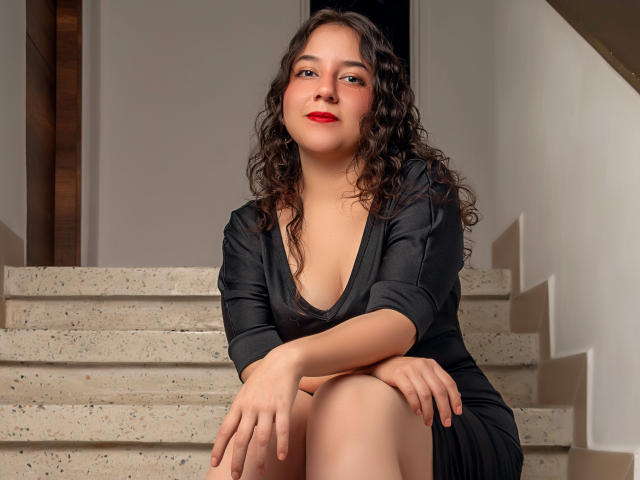 ChloeVelvet with curly brown hair, red lipstick, and a black dress, sitting on stone stairs, knees up, hands resting on knees, smiling slightly, showing her small tits.