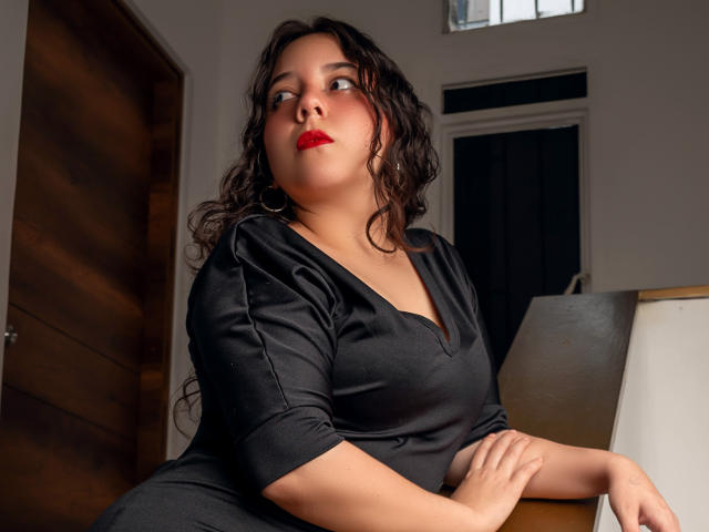 ChloeVelvet has curly black hair, red lipstick, and wears a black dress, sitting with hands on a table, looking to the side, with wooden door in background.