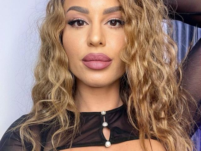 YourWetKate-hot with wavy blonde hair, full pink lips, and brown eyes, wearing a sheer black top with small pearl buttons, looking straight at the camera.