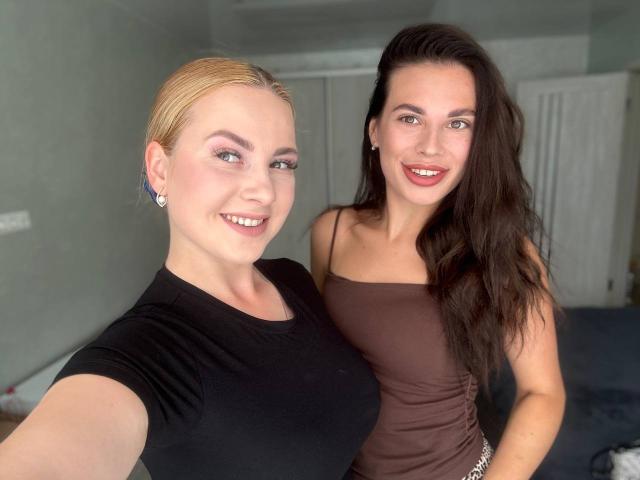 GlossyTwins with blonde hair and black top, tits visible, standing next to GlossyTwins with brown hair and brown top, both smiling, in a room with white curtains.