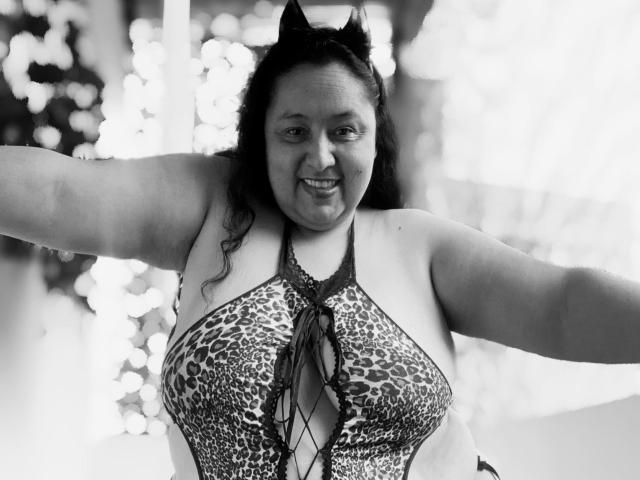 A TistMature tem seios grandes e redondos, vestindo um top de alças de leopardo com um fecho frontal de renda, orelhas de gato, e os braços abertos, sorrindo em uma foto em preto e branco..