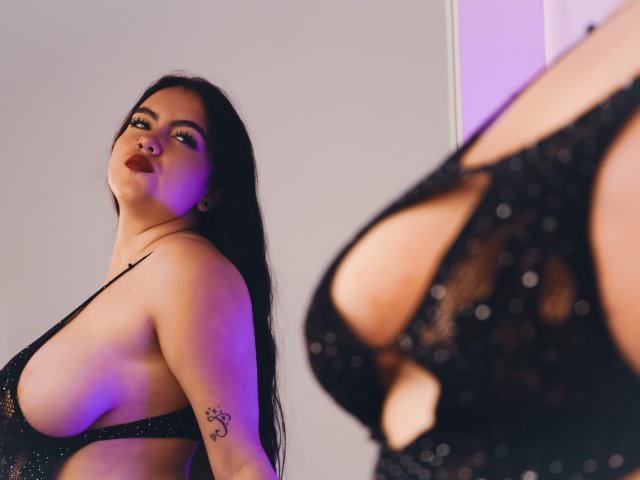 ZoeQuinn with long black hair, red lipstick, and a tattoo on her arm, wearing a sparkly black lace-up dress, showing her big tits in a mirror reflection.