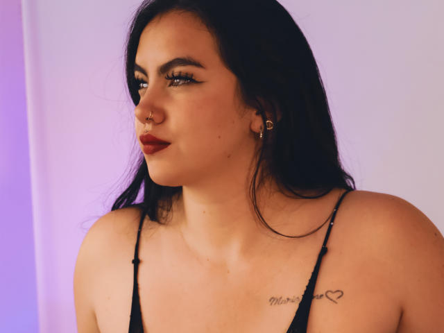 ZoeQuinn with dark hair, red lipstick, and black eyeliner, wearing a black spaghetti strap top, nose ring, gold hoop earrings, tattoo on right shoulder, smiling.