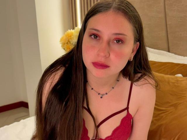 WidnyBeaumont with long brown hair, red makeup, and red lace top, showing small tits, wearing a bead necklace, sitting on a bed with yellow pillows.