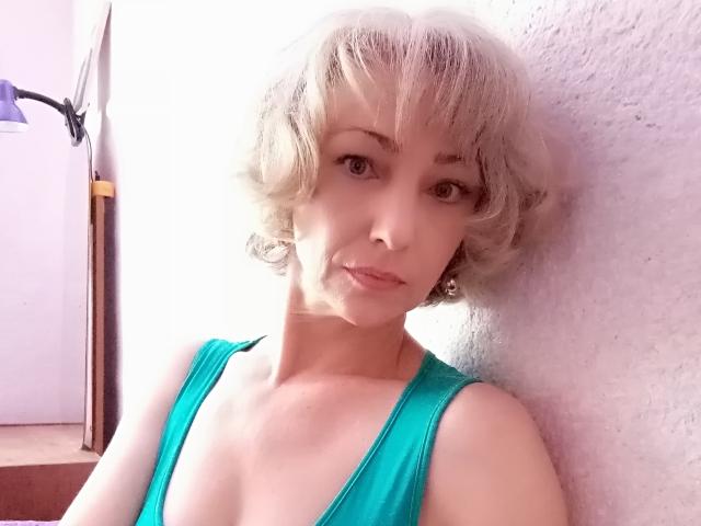 LorieCarrieFleur with short, curly, blonde hair wearing a teal, low-cut top, showing her small tits and fair skin, leaning against a white wall, looking at the camera.