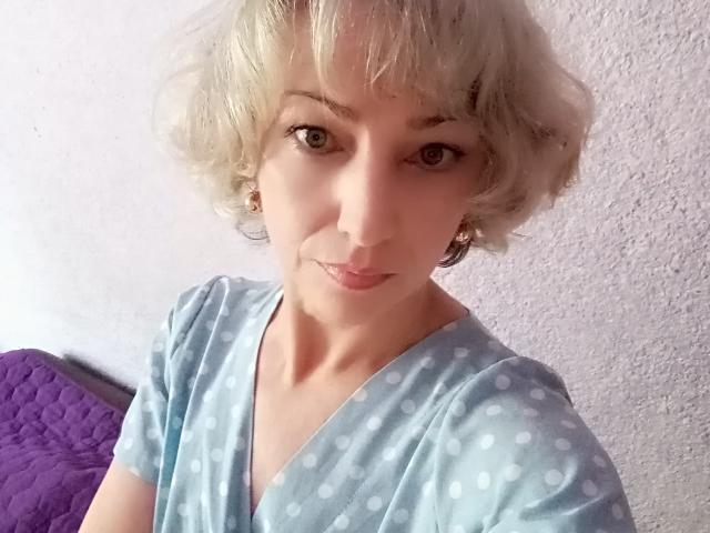 LorieCarrieFleur has short, curly blonde hair, light skin, and wears a blue polka dot shirt. She's smiling with small tits, purple quilted pillow in the background.