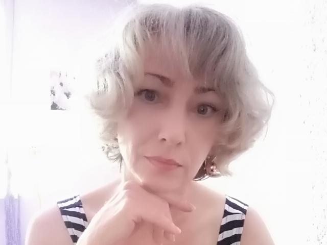 LorieCarrieFleur with short, wavy grayish-blonde hair, wearing a black-and-white striped tank top, resting her chin on her hand, looking at the camera with a slight smile.