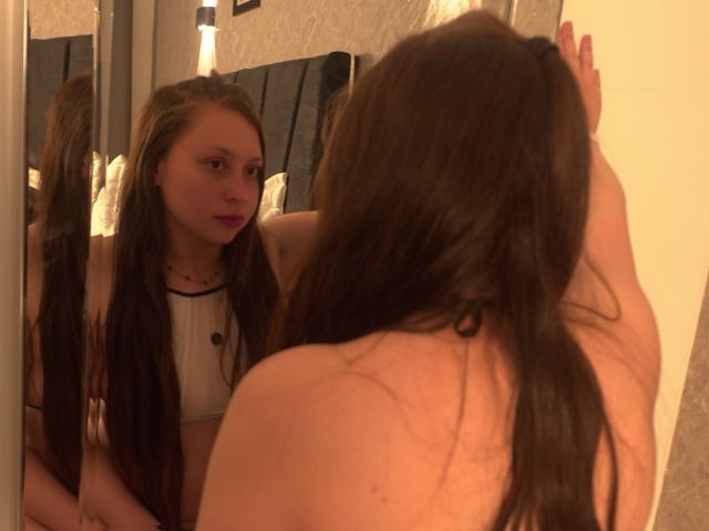 WidnyBeaumont with long brown hair, looking at herself in a mirror, showing her bare back and small tits, wearing a white top, in a dimly lit room.
