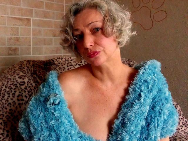 LorieCarrieFleur with short curly gray hair, wearing a fluffy blue top, exposing her boobs, sitting on a leopard print couch, winking, with pink lipstick.