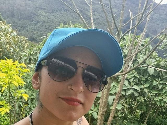 XimenaNahomi with a blue cap, nose ring, and dark sunglasses, smiling with red lipstick, green leafy background, and a tree behind her.