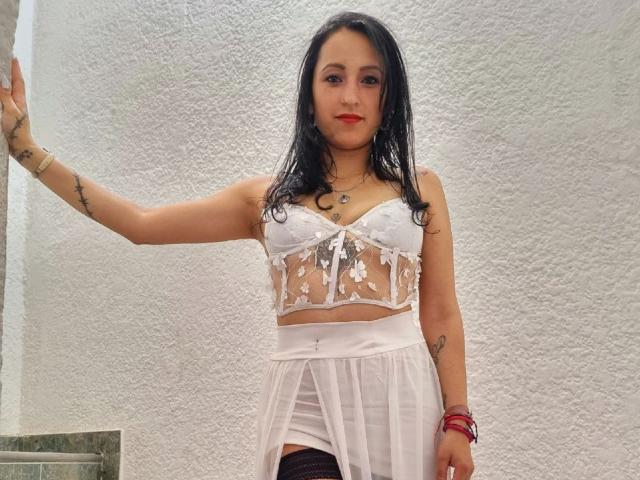XimenaNahomi with wet black hair, wearing a sheer white bralette and skirt, showing small tits and a visible gape in her skirt, standing against a textured white wall.