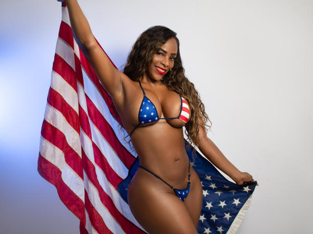 Isabella, con i capelli castani ricci, rossetto rosso, indossa un bikini blu e rosso con motivo a stelle, tiene in mano una grande bandiera americana, mostrando il suo seno prosperoso e uno slip minuscolo, sorridendo..
