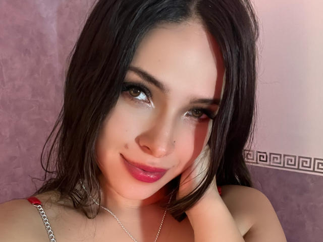 LorreineMontclair has shiny brown eyes, dark wet hair, pink lipstick, and a nose ring. She's smiling with a hand on her cheek, wearing a red strap and silver necklace.