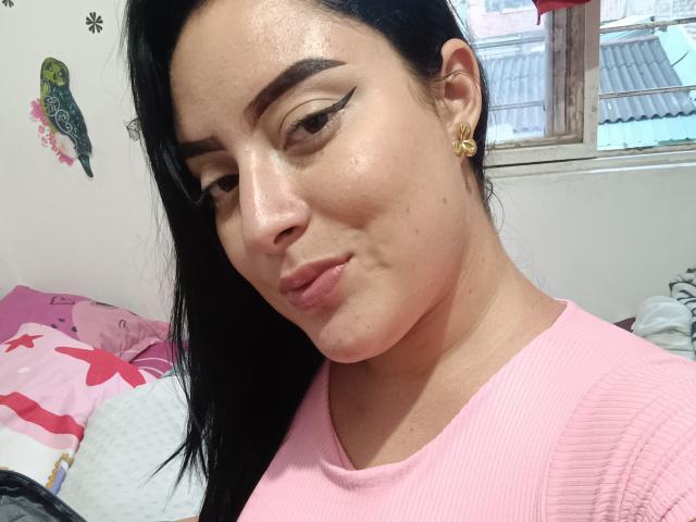 Gyar has glossy skin, black eyebrows, and winged eyeliner, wearing a pink ribbed shirt, with a small gold earring, pouting slightly, and showing a cute, subtle smile.