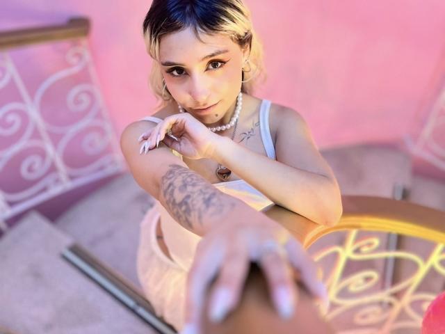 MaveParker with blonde hair, white tank top, and pearl necklace, reaching forward with tattooed arm, pink-lit background, white metal railing, and wooden chair in foreground.