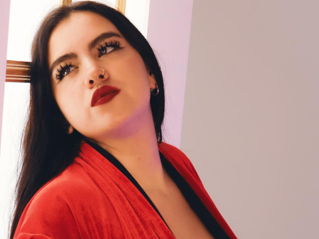 ZoeQuinn with long black hair, red lipstick, and nose ring, wearing a red robe, looking up with heavy eyelashes, soft pink light on her cheek.