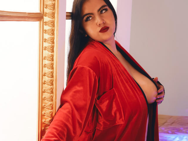 ZoeQuinn with long black hair, red satin robe, deep cleavage, showing her big tits, red lipstick, and a nose ring, standing by a ornate window.