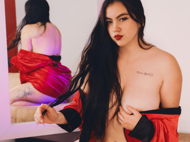ZoeQuinn with long black hair, red lipstick, and a red satin robe, exposing her tits, holding her hair, tattoo on right chest, tattoo on left thigh, mirror reflection.