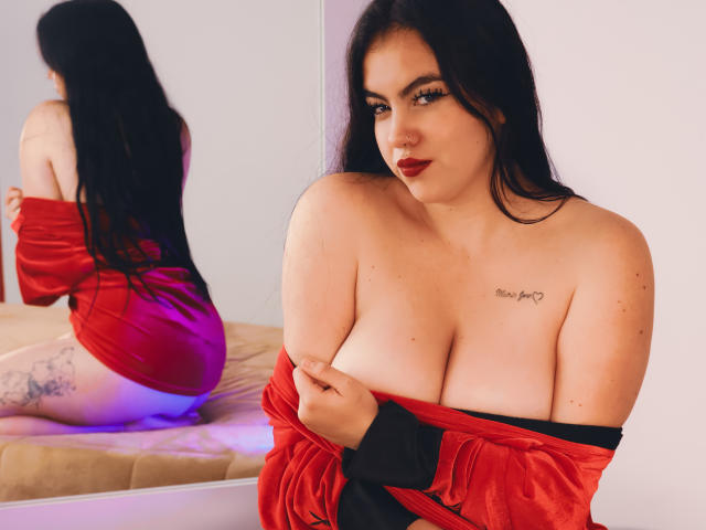 ZoeQuinn with long black hair, red velvet dress pulled down to expose large tits, red lipstick, tattoo on right chest, sitting on bed, reflection in mirror shows her ass.