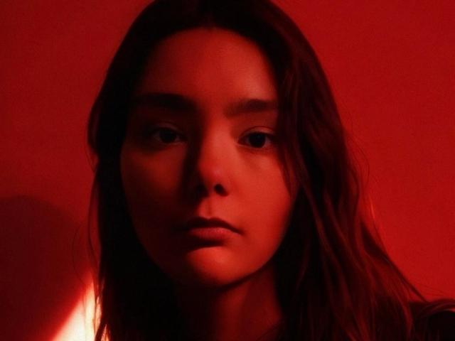 NyxSari has red-tinted skin, dark wet hair, and a serious expression, with a small warm light on the left. Her face is centered, and she looks straight at the camera.
