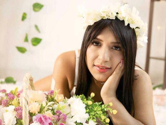 IsabellaStonnee with long dark hair, white flower crown, smiling, hand on cheek, lying on bed, holding bouquet of pink, white, and yellow flowers, wearing white dress.