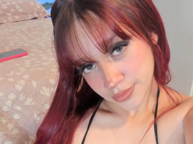 AdaFerreira with red hair, bangs, and black eyeliner, smiling, wearing a black halter top, showing her tits, lying on a bed with patterned sheets, smartphone visible.
