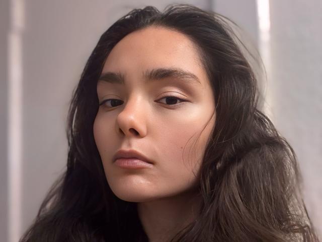 NyxSari has wavy dark brown hair, a light tan skin tone, and a serious expression with subtle makeup, showing her nose and full lips. Her eyebrows are naturally thick.