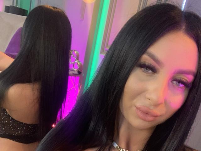 LexyVoss has long black hair, full eyebrows, and wears a glittery black bra. Her reflection shows her back. She has a nose ring and a pouty expression.