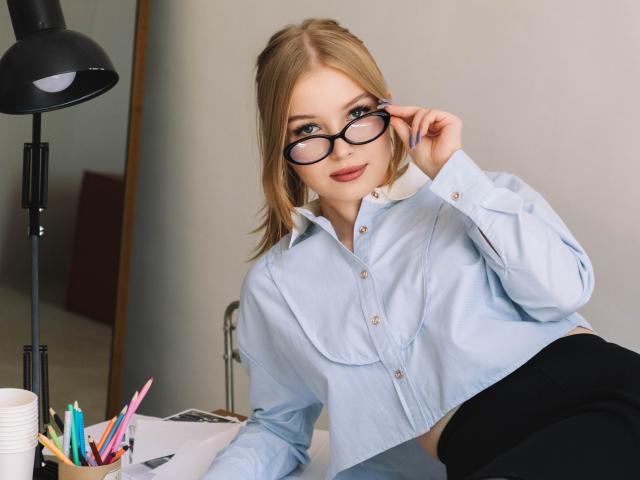 NicoleBrian with blonde hair, blue eyes, wearing glasses, light blue shirt, black skirt, sitting at desk, holding glasses, smiling, black lamp, colorful pencils, white cup.