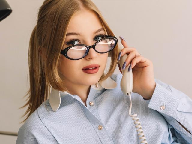 NicoleBrian with blonde hair, wearing glasses, light blue shirt with white collar, holding a white corded phone, has purple nail polish, and is looking into the camera.